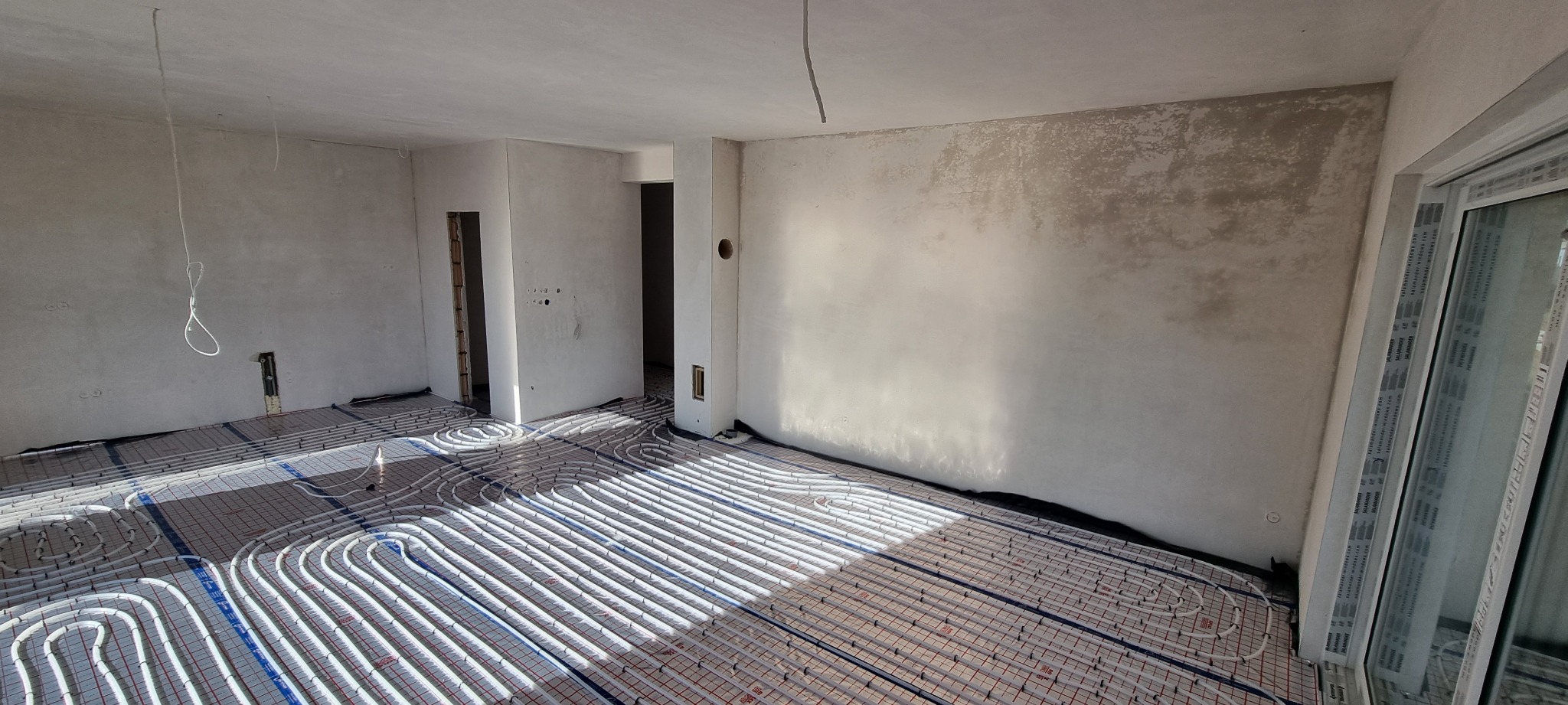 Rozłożone rury ogrzewania podłogowego na izolacji w surowym pomieszczeniu, widoczne ściany w trakcie wykończenia i okno balkonowe.