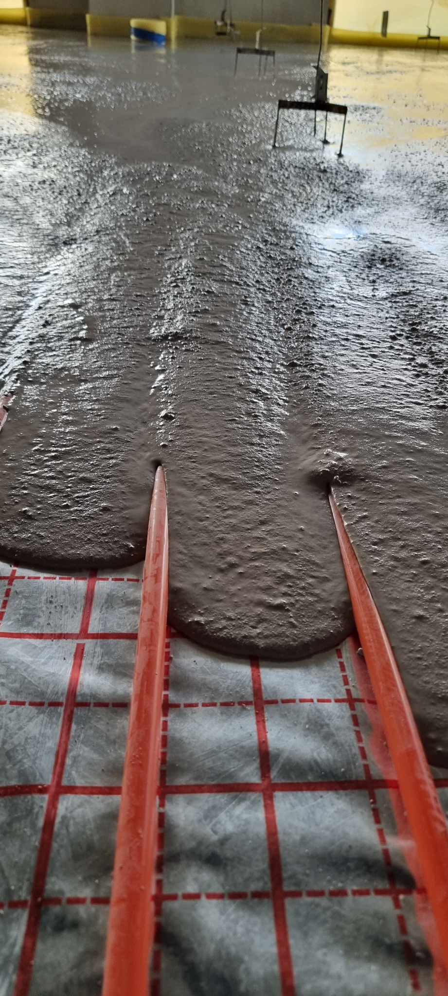 Wylewka samopoziomująca rozprowadzana na rurach ogrzewania podłogowego ułożonych na folii z czerwoną siatką, widoczne narzędzia do rozprowadzania w tle.