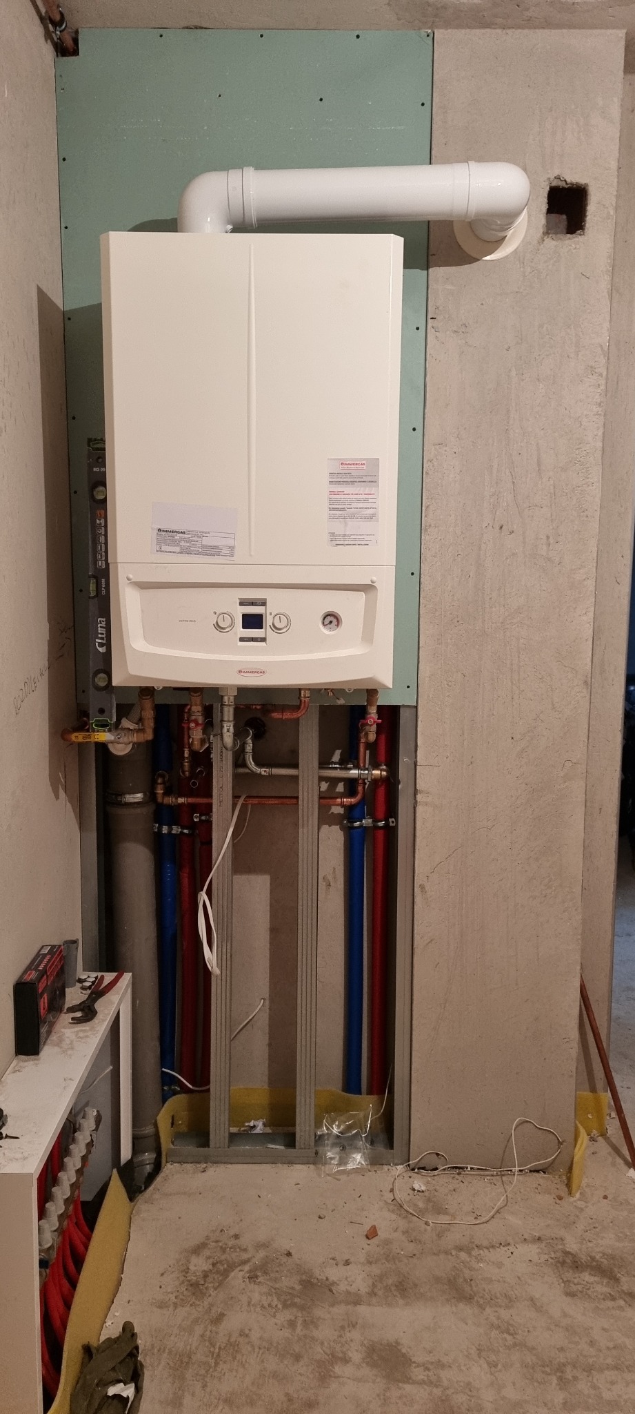 Biały piec gazowy marki Immergas zamontowany na ścianie z płyt kartonowo-gipsowych, widoczne rury miedziane i plastikowe, instalacja w trakcie prac wykończeniowych.