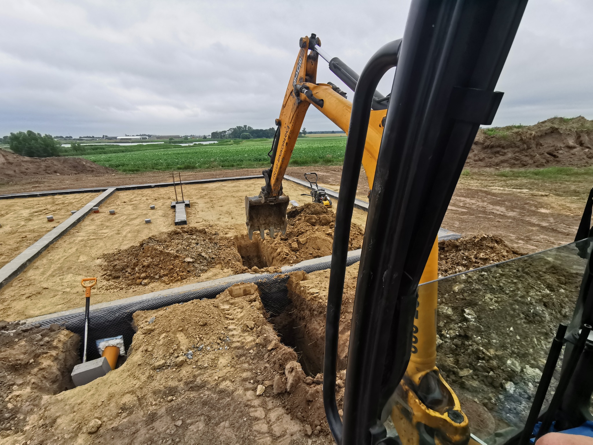 Żółta koparka wykopuje rów pod instalację na placu budowy z widokiem na pola, widoczne elementy instalacji, szpadel i zagęszczarka w tle.