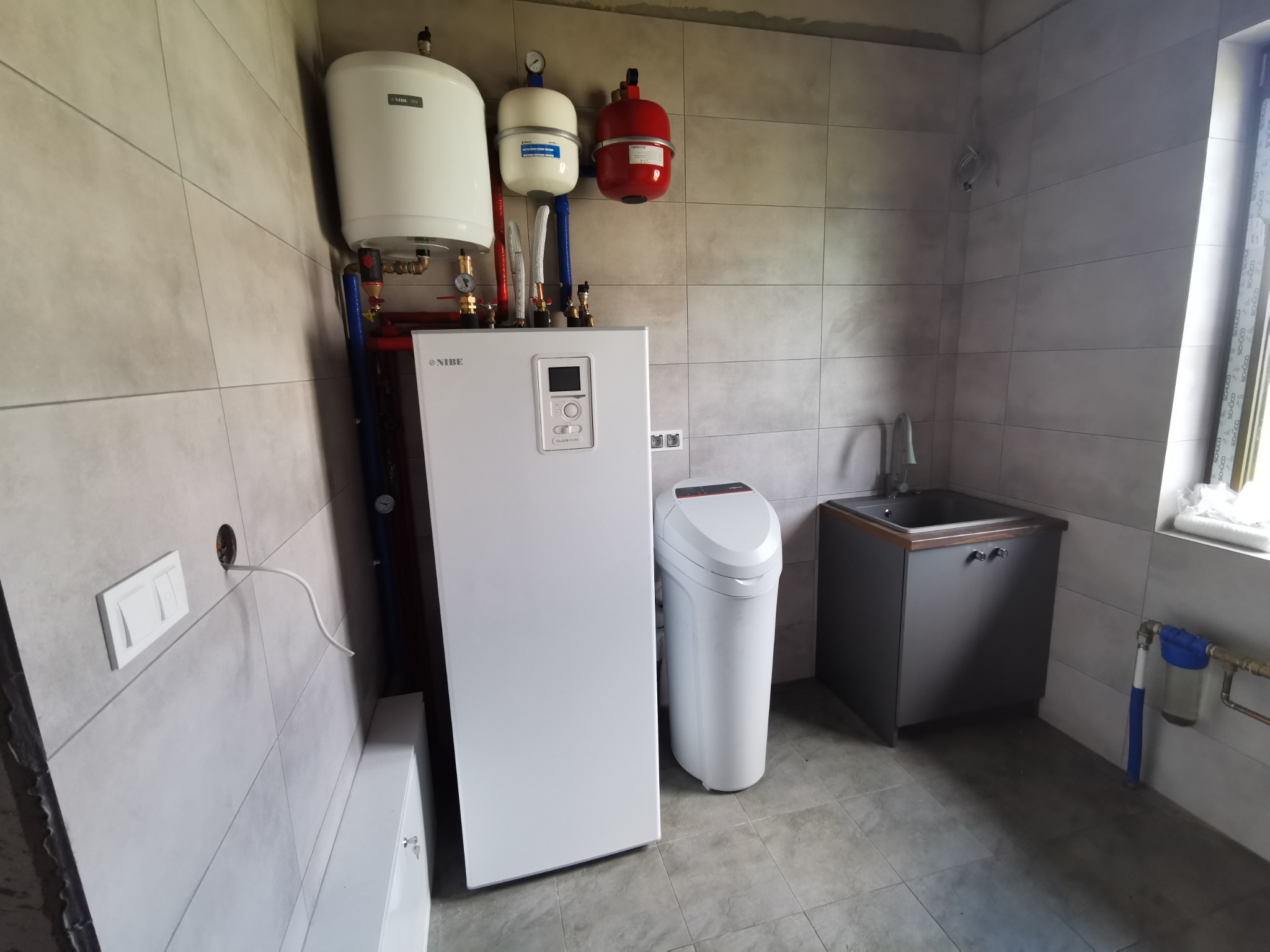 Biały piec grzewczy NIBE z podłączonymi zbiornikami wyrównawczymi i zmiękczaczem wody w pomieszczeniu wyłożonym szarymi płytkami ceramicznymi.