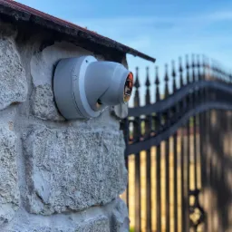 Kamera monitoringu kopułkowa, zamontowana na kamiennym słupku ogrodzenia, z kutą bramą w tle i błękitnym niebem