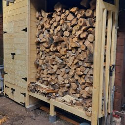 SAWA Agata Woroniecka-Mrozińska - Drewniana wiata na drewno z zamkniętą szafką, wypełniona pociętymi klockami drewna. Na ziemi leżą narzędzia ogrodnicze i wiaderko.