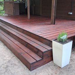 SAWA Agata Woroniecka-Mrozińska - Ciemnobrązowy taras z drewna ze schodami, donica z ziołami i dekoracją w kształcie ptaka. Tło: drewniany budynek i zieleń. Widok z dołu.