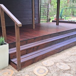 SAWA Agata Woroniecka-Mrozińska - Ciemnobrązowy taras drewniany z balustradą i schodami, z donicą z lawendą. Widok na ogród z huśtawką. Deski tarasowe lakierowane, z widoczną strukturą drewna.