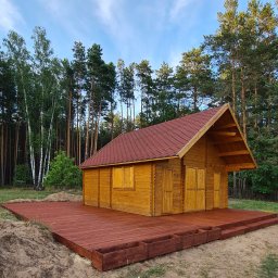 SAWA Agata Woroniecka-Mrozińska - Drewniany domek z czerwonym dachem na drewnianym tarasie otoczony lasem. Widoczne detale konstrukcji i naturalne otoczenie. Słoneczny dzień.