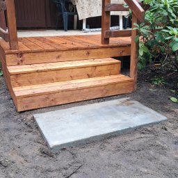 SAWA Agata Woroniecka-Mrozińska - Drewniane schody tarasowe z balustradą, prowadzące do brązowego budynku, z betonową płytą u podstawy, otoczone ziemią i zielenią. Widok z dołu.