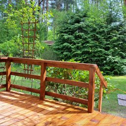 SAWA Agata Woroniecka-Mrozińska - Błyszczący, drewniany taras z brązową balustradą, widok na zielony ogród z dywanem i dekoracyjną kratką. Świeże drewno w ciepłym oświetleniu.