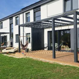 Nowoczesny taras drewniany z czarną pergolą i hamakiem, przylegający do białego domu z dużymi oknami, otoczony zielonym trawnikiem i młodymi nasadzeniami.