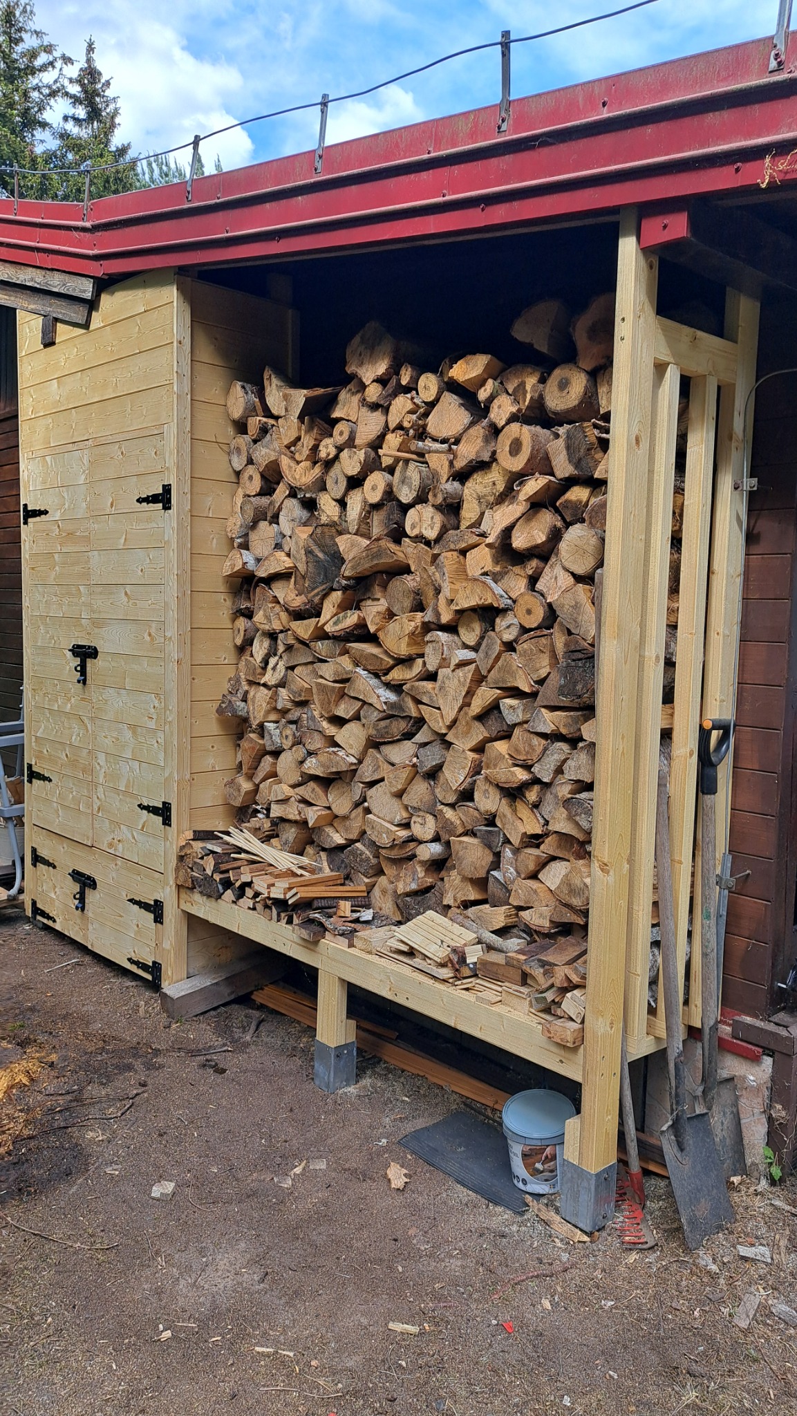 Drewniana wiata na drewno z zamkniętą szafką, wypełniona pociętymi klockami drewna. Na ziemi leżą narzędzia ogrodnicze i wiaderko.