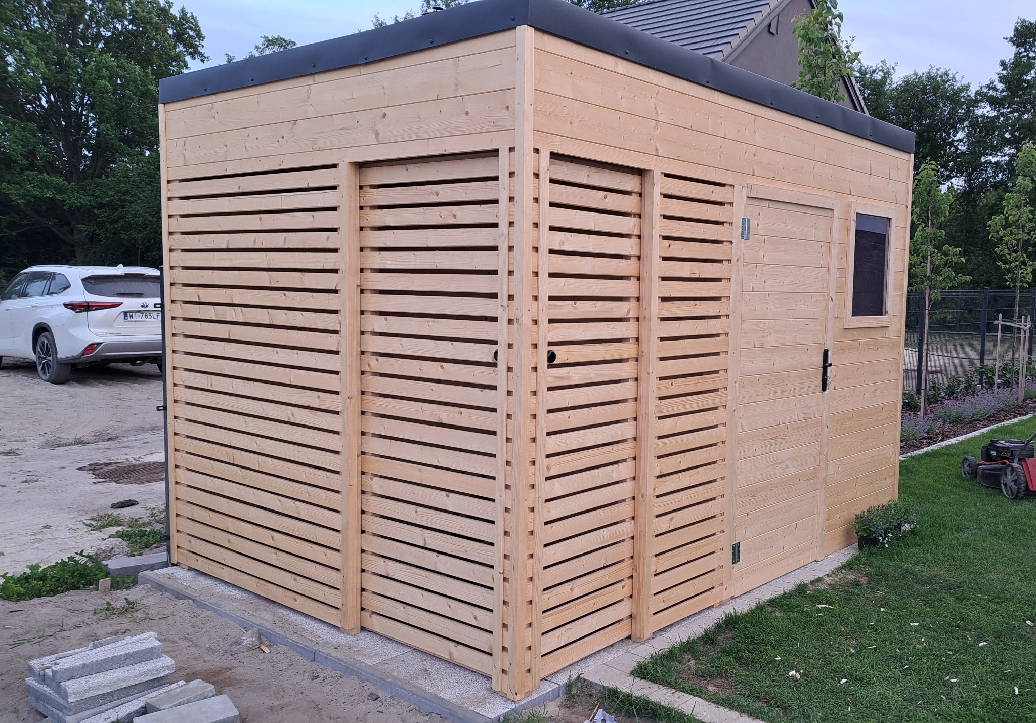 Nowoczesna, drewniana konstrukcja z poziomymi listwami i małym oknem, prawdopodobnie domek narzędziowy, na tle zieleni i samochodu.