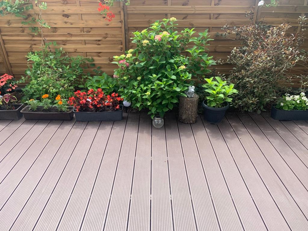 Jasny taras z desek kompozytowych, otoczony różnorodną zielenią w donicach i drewnianym płotem. Dekoracje: szklany lampion i rzeźba myszy na pniu.