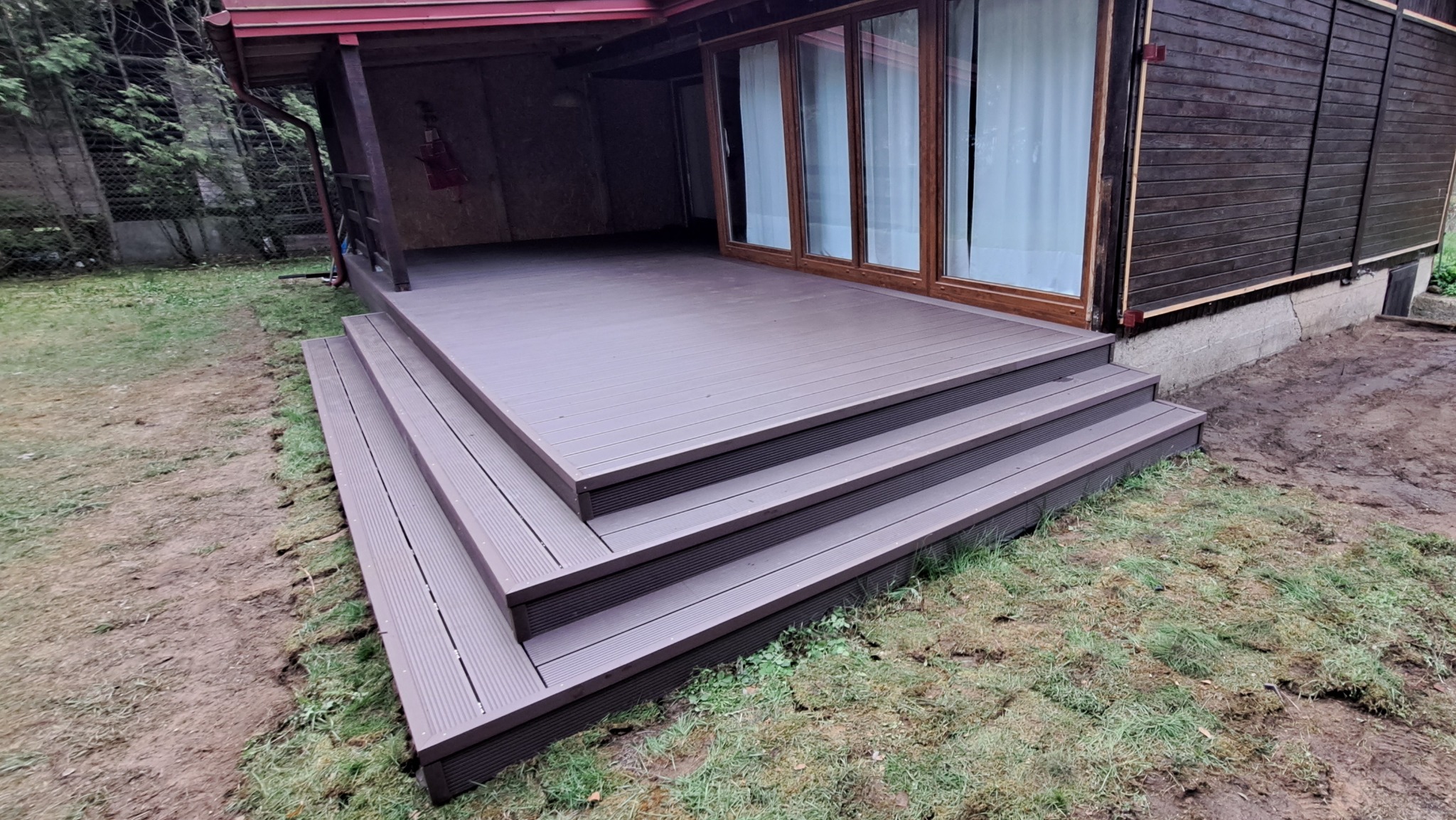 Brązowy taras kompozytowy z trzema schodkami, przylegający do budynku z oknami i ciemną elewacją. Widoczne połączenia desek i trawa wokół.