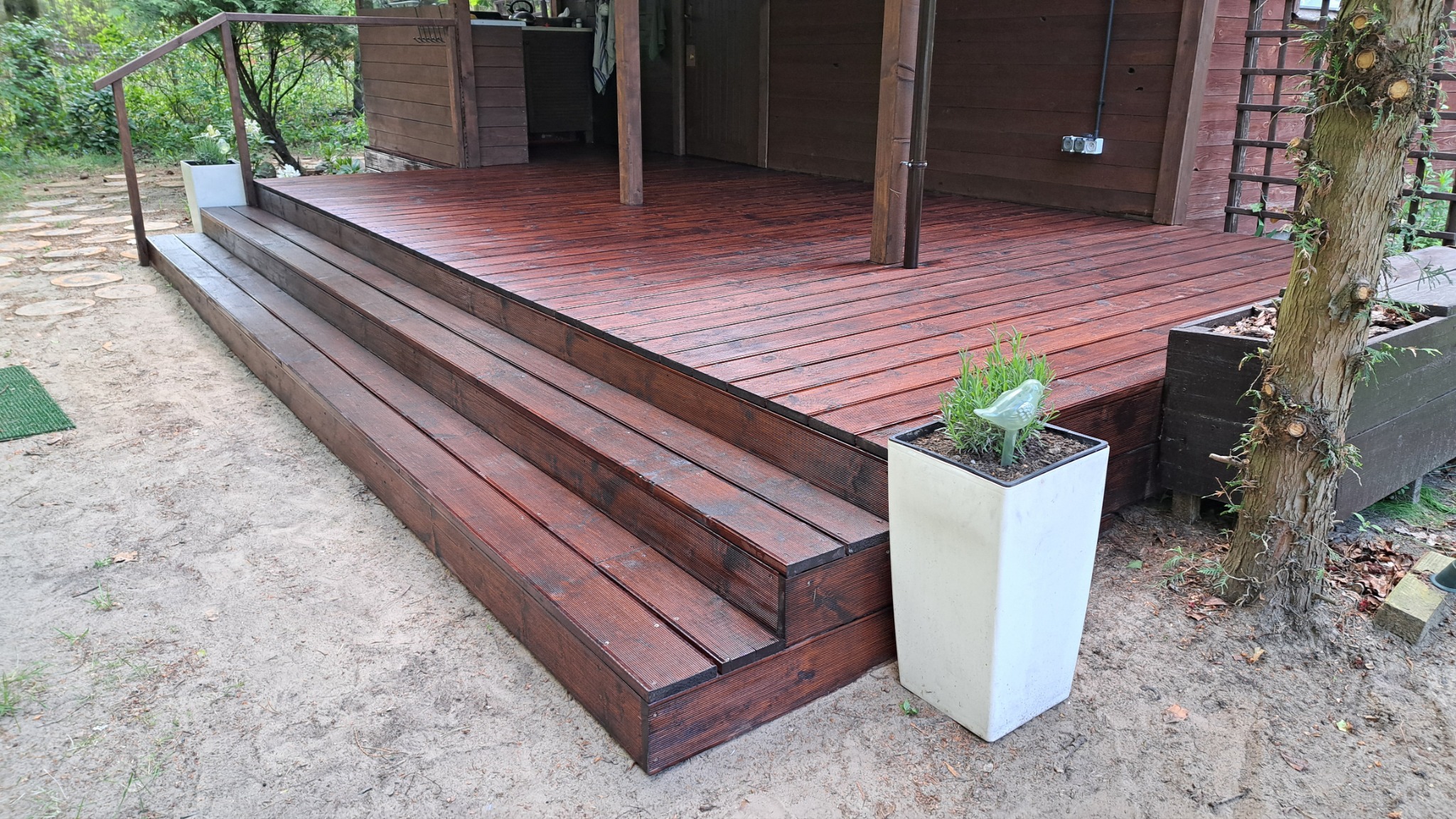 Ciemnobrązowy taras z drewna ze schodami, donica z ziołami i dekoracją w kształcie ptaka. Tło: drewniany budynek i zieleń. Widok z dołu.
