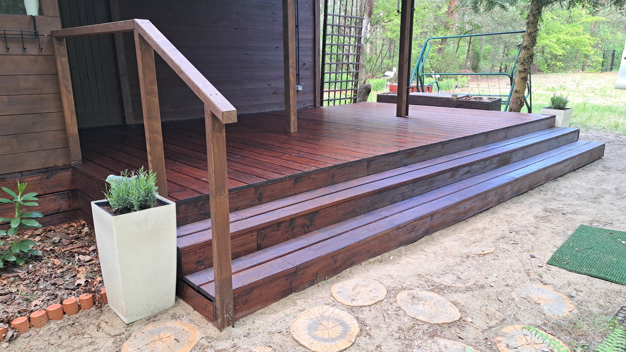 Ciemnobrązowy taras drewniany z balustradą i schodami, z donicą z lawendą. Widok na ogród z huśtawką. Deski tarasowe lakierowane, z widoczną strukturą drewna.