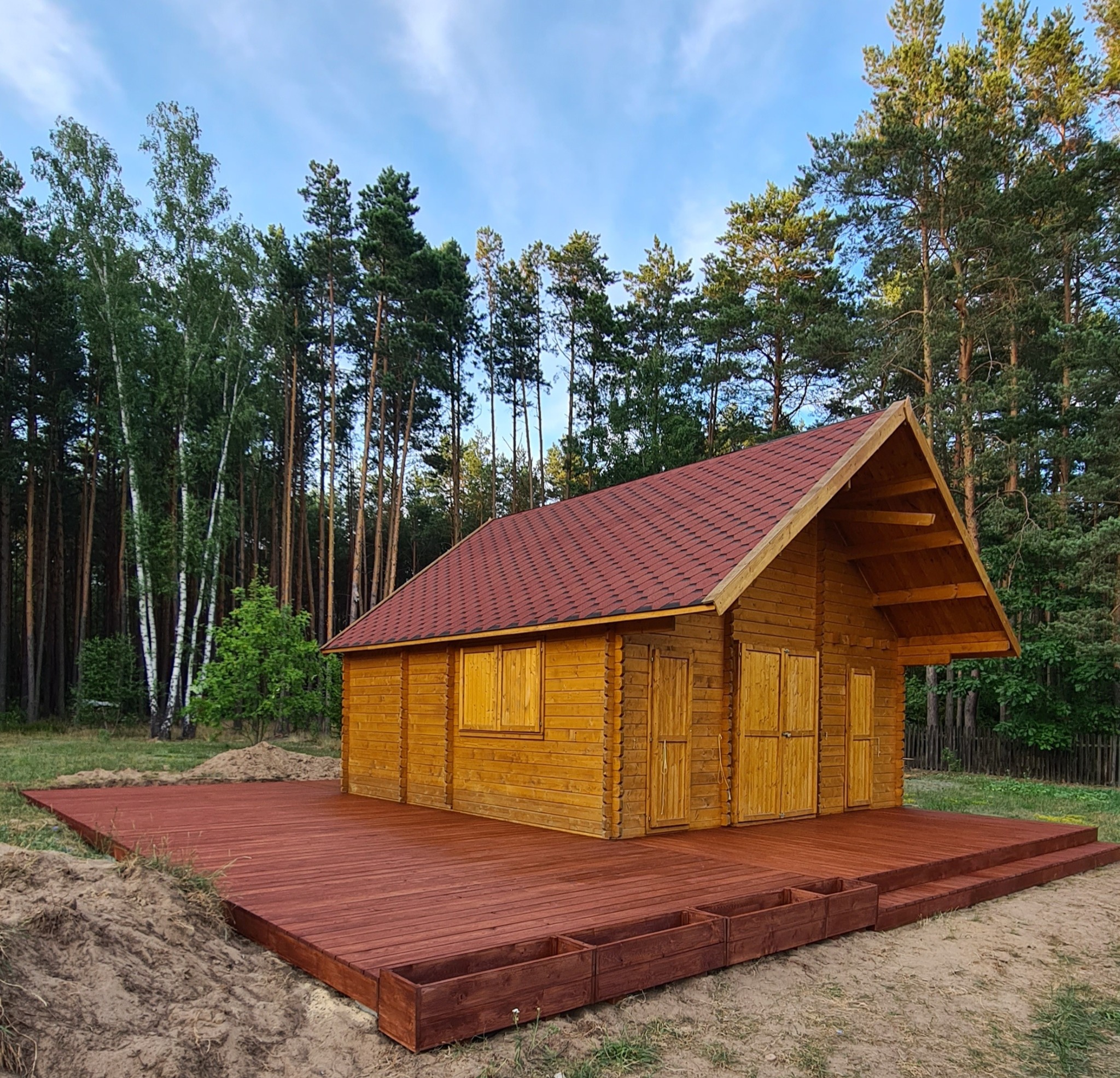 Drewniany domek z czerwonym dachem na drewnianym tarasie otoczony lasem. Widoczne detale konstrukcji i naturalne otoczenie. Słoneczny dzień.