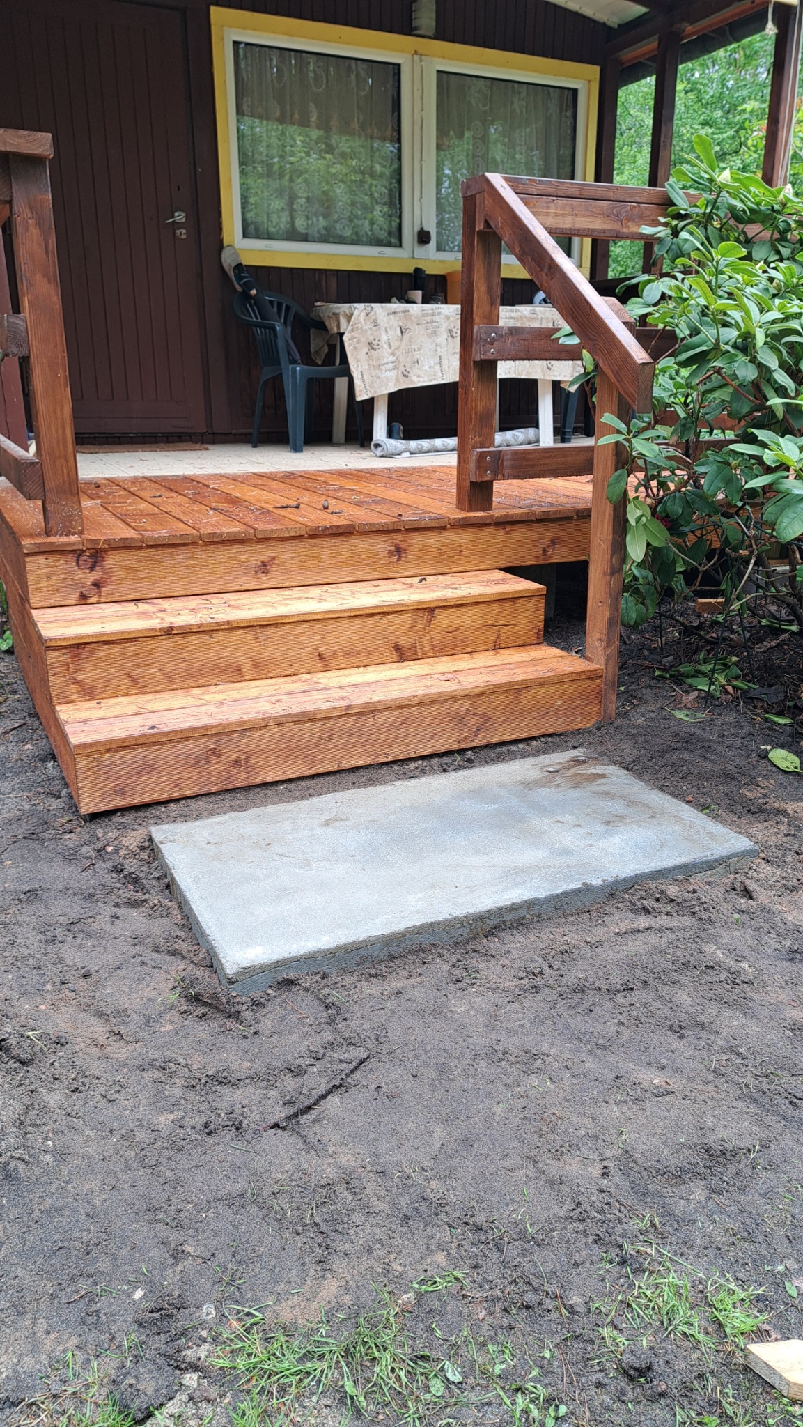 Drewniane schody tarasowe z balustradą, prowadzące do brązowego budynku, z betonową płytą u podstawy, otoczone ziemią i zielenią. Widok z dołu.