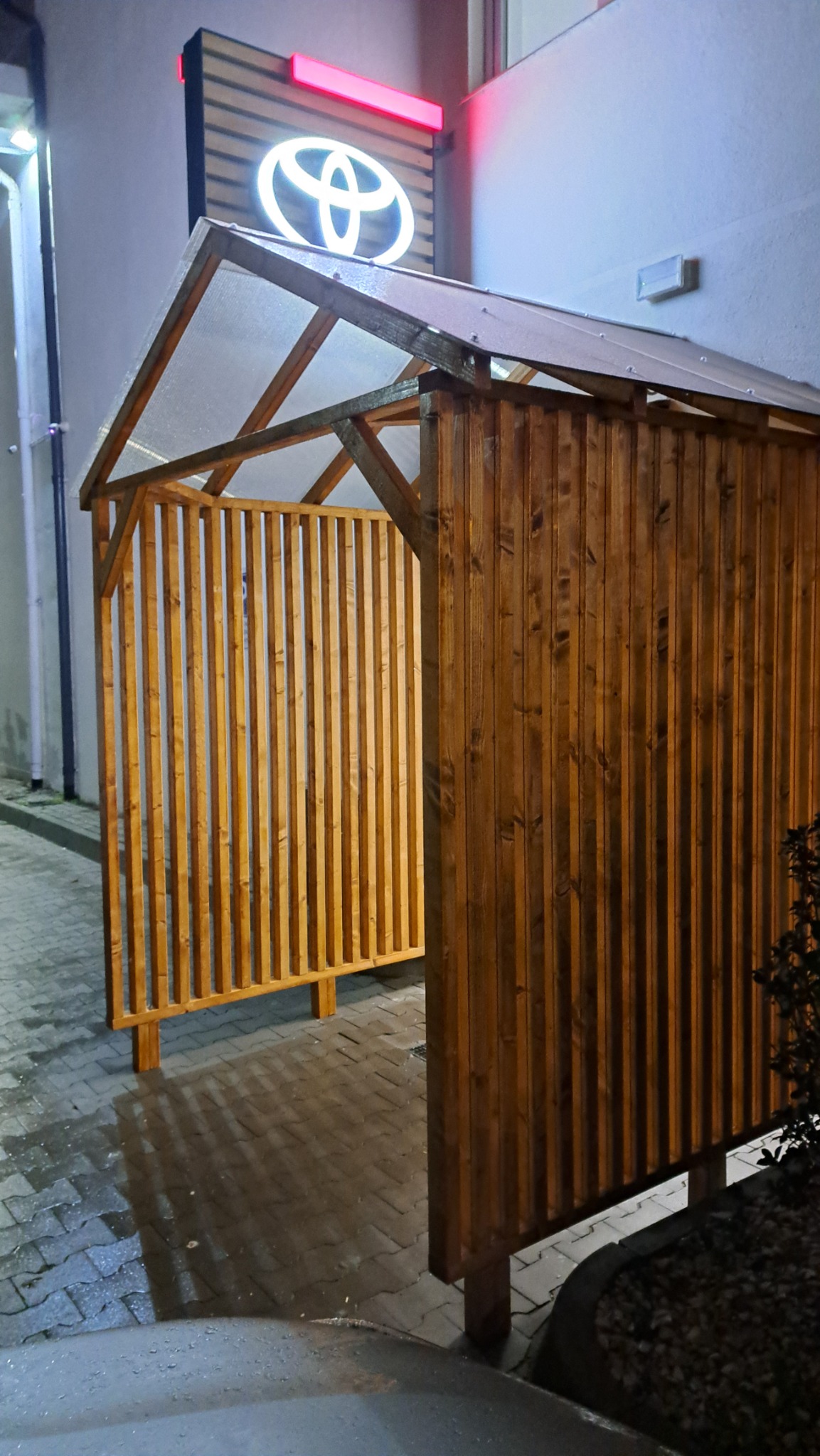 Drewniana pergola z przezroczystym dachem, oświetlona nocą, z logo Toyota w tle. Konstrukcja z pionowych desek w ciepłym odcieniu brązu.