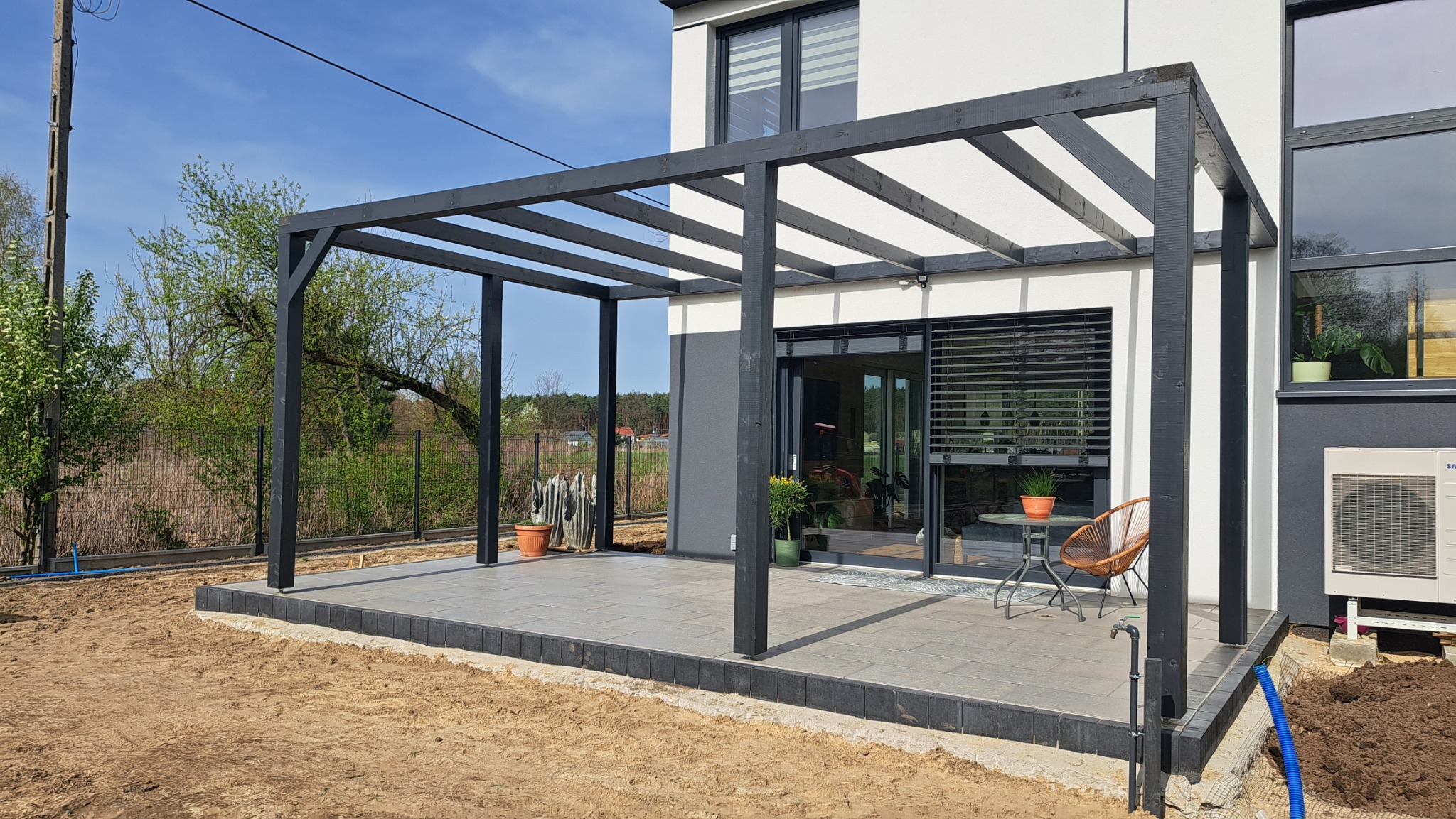 Nowoczesna pergola z ciemnego drewna na tarasie z szarych płyt. Widok na dom z dużymi oknami i zadbany ogród. Stolik z krzesłem na tarasie.