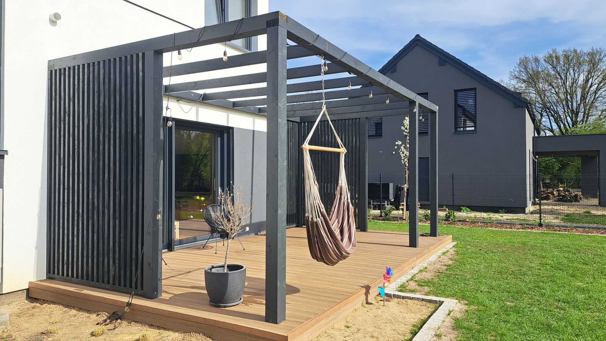 Nowoczesna pergola z ciemnego drewna na jasnym tarasie, hamak w środku. Dom w tle. Elementy dekoracyjne jak lampki i wiatraczek dodają uroku.