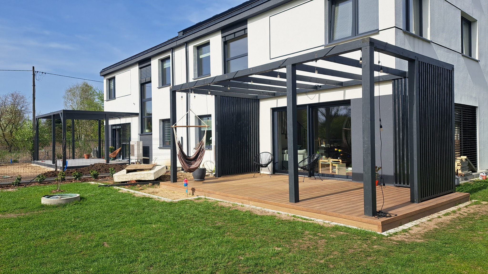 Nowoczesny taras drewniany z czarną pergolą i hamakiem, przylegający do białego domu z dużymi oknami, otoczony zielonym trawnikiem i młodymi nasadzeniami.