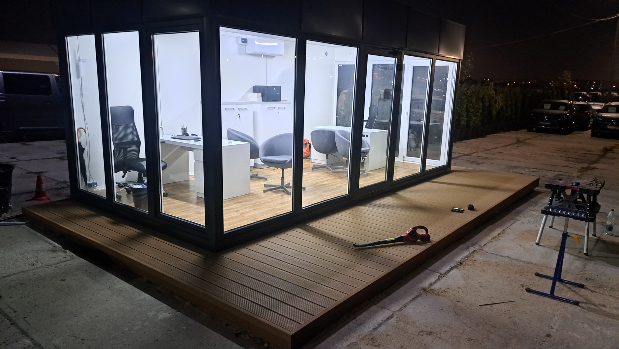 Nowoczesna, przeszklona kabina biurowa z tarasem. Widoczne biurka, krzesła i klimatyzacja wewnątrz. Na zewnątrz narzędzia i oświetlenie nocne.