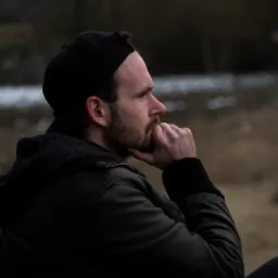 Mężczyzna z brodą, w czapce i kurtce, zamyślony, profil, ręka pod brodą, w tle krzyż na wzgórzu.