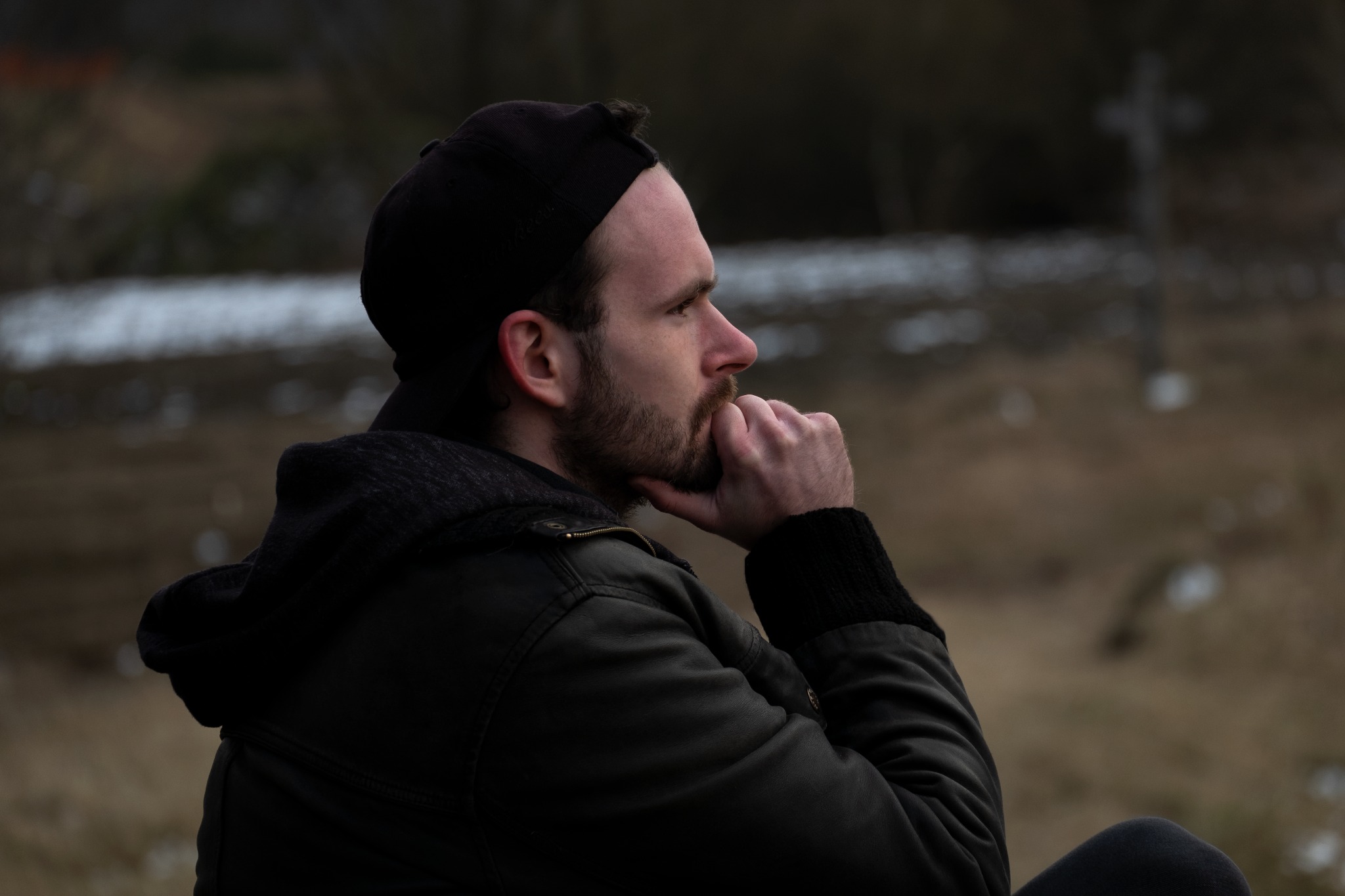 Mężczyzna z brodą, w czapce i kurtce, zamyślony, profil, ręka pod brodą, w tle krzyż na wzgórzu.