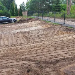 Wyrównany, wilgotny teren budowlany z widocznymi śladami opon i równiarki, ogrodzony siatką, z zaparkowanym czarnym kombi w tle i budynkiem mieszkalnym.