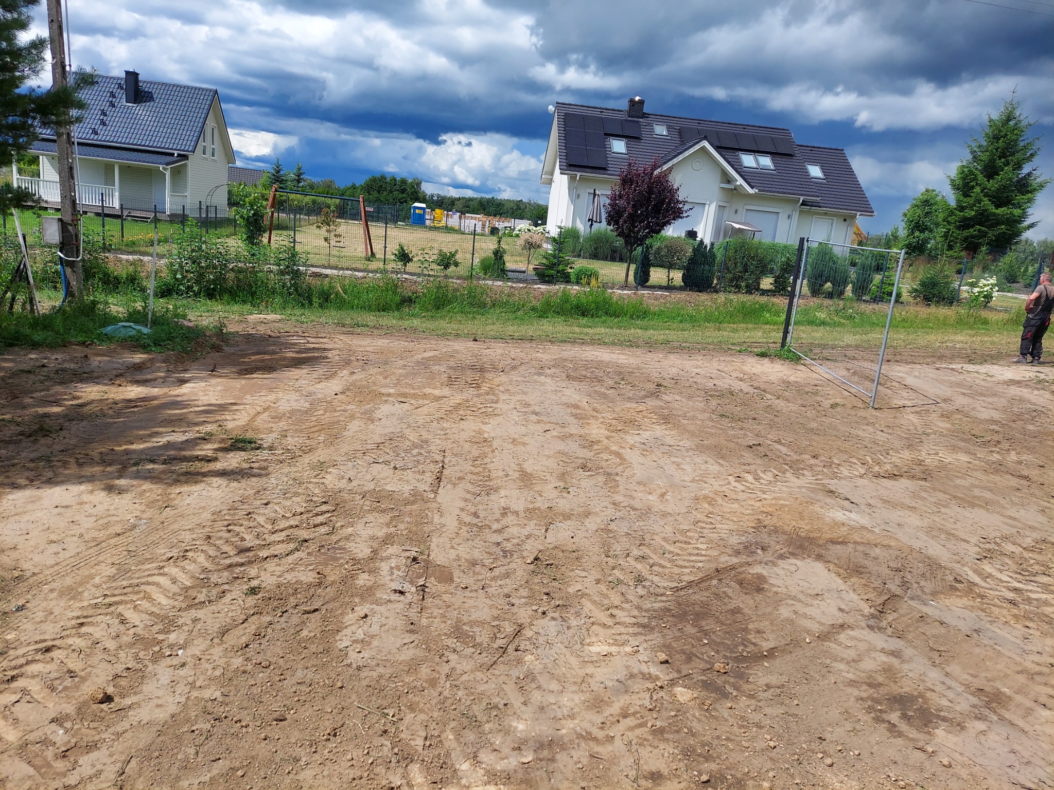 Przygotowany teren pod budowę domu jednorodzinnego, widoczne ślady opon maszyn budowlanych na ubitej ziemi, w tle drugi dom, ogrodzenie i pochmurne niebo.