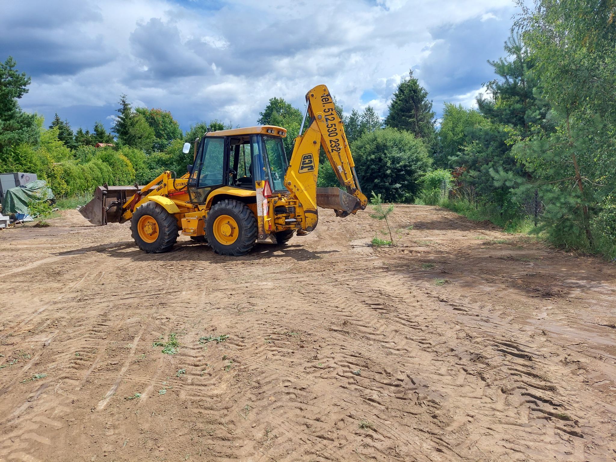 Żółta koparko-ładowarka JCB na placu budowy z widocznymi śladami opon w gruncie, zieleń w tle i numer telefonu na ramieniu koparki.