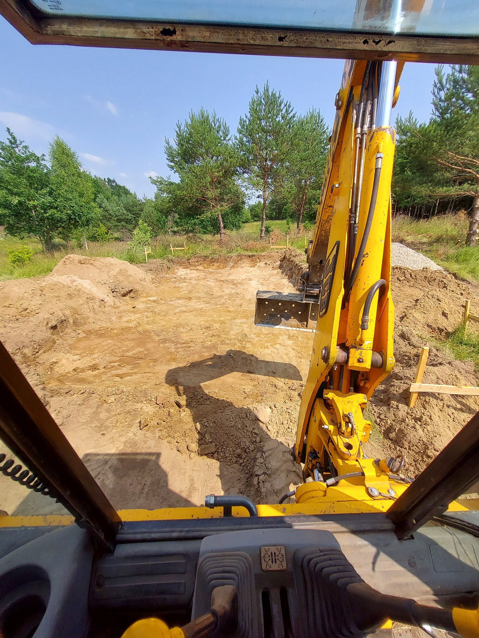 Widok z kabiny żółtej koparki na wykop pod fundamenty z widocznymi drewnianymi szalunkami i stertą żwiru w tle.