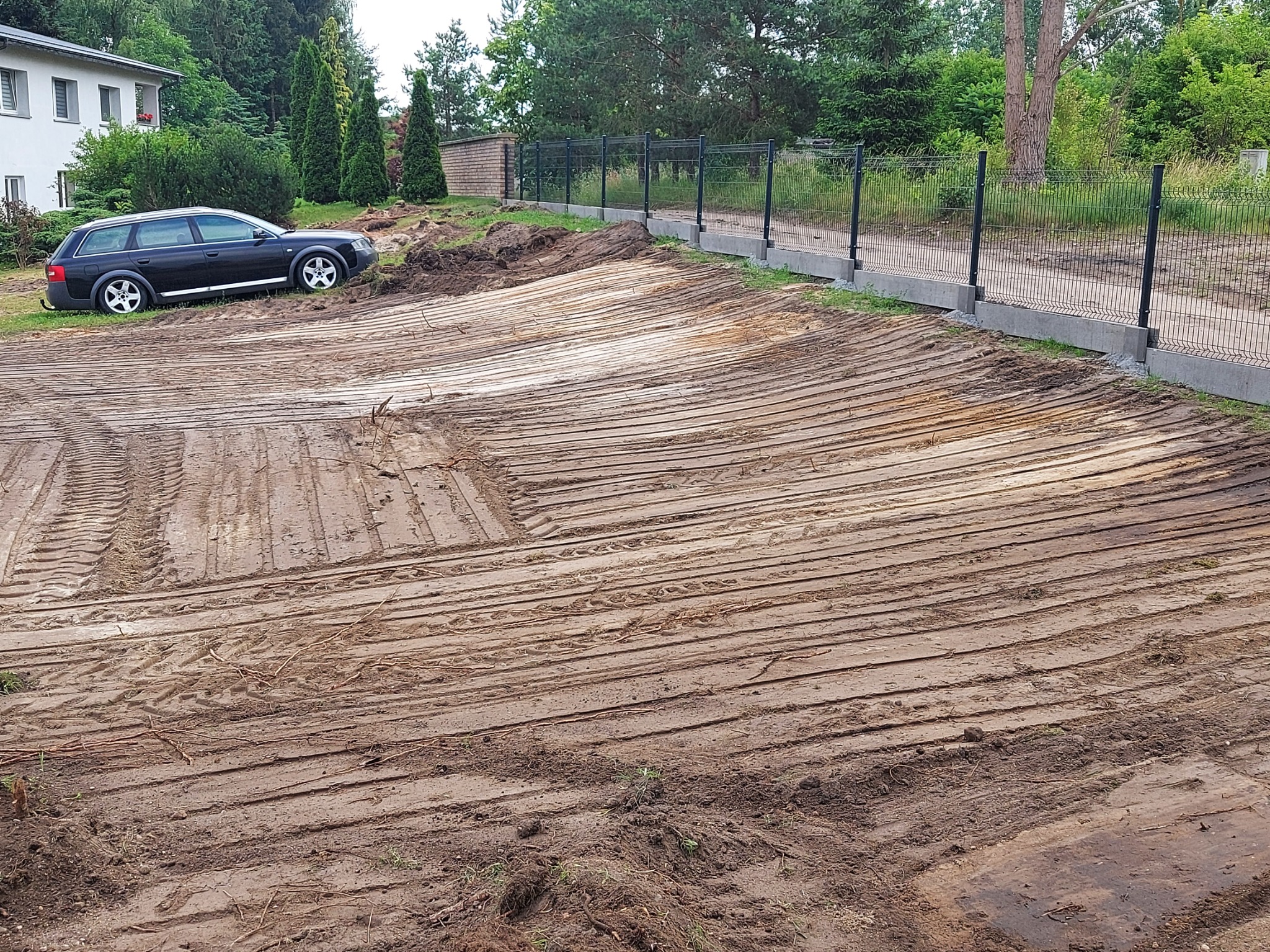 Wyrównany, wilgotny teren budowlany z widocznymi śladami opon i równiarki, ogrodzony siatką, z zaparkowanym czarnym kombi w tle i budynkiem mieszkalnym.