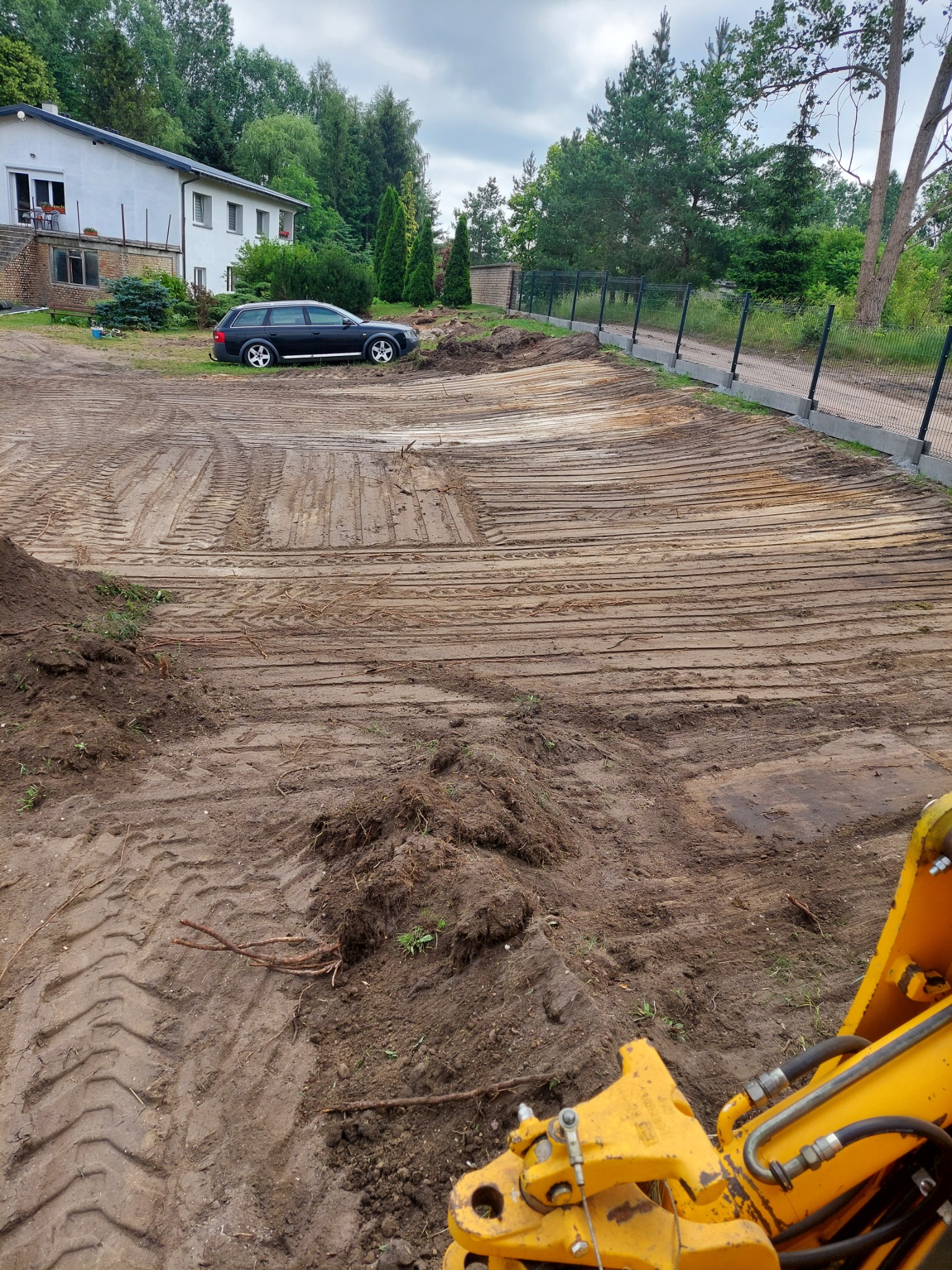 Przygotowany teren pod budowę, widoczne ślady po maszynach, częściowo wyrównana powierzchnia, w tle dom jednorodzinny i zaparkowany samochód osobowy, z prawej strony fragment żółtej koparki.