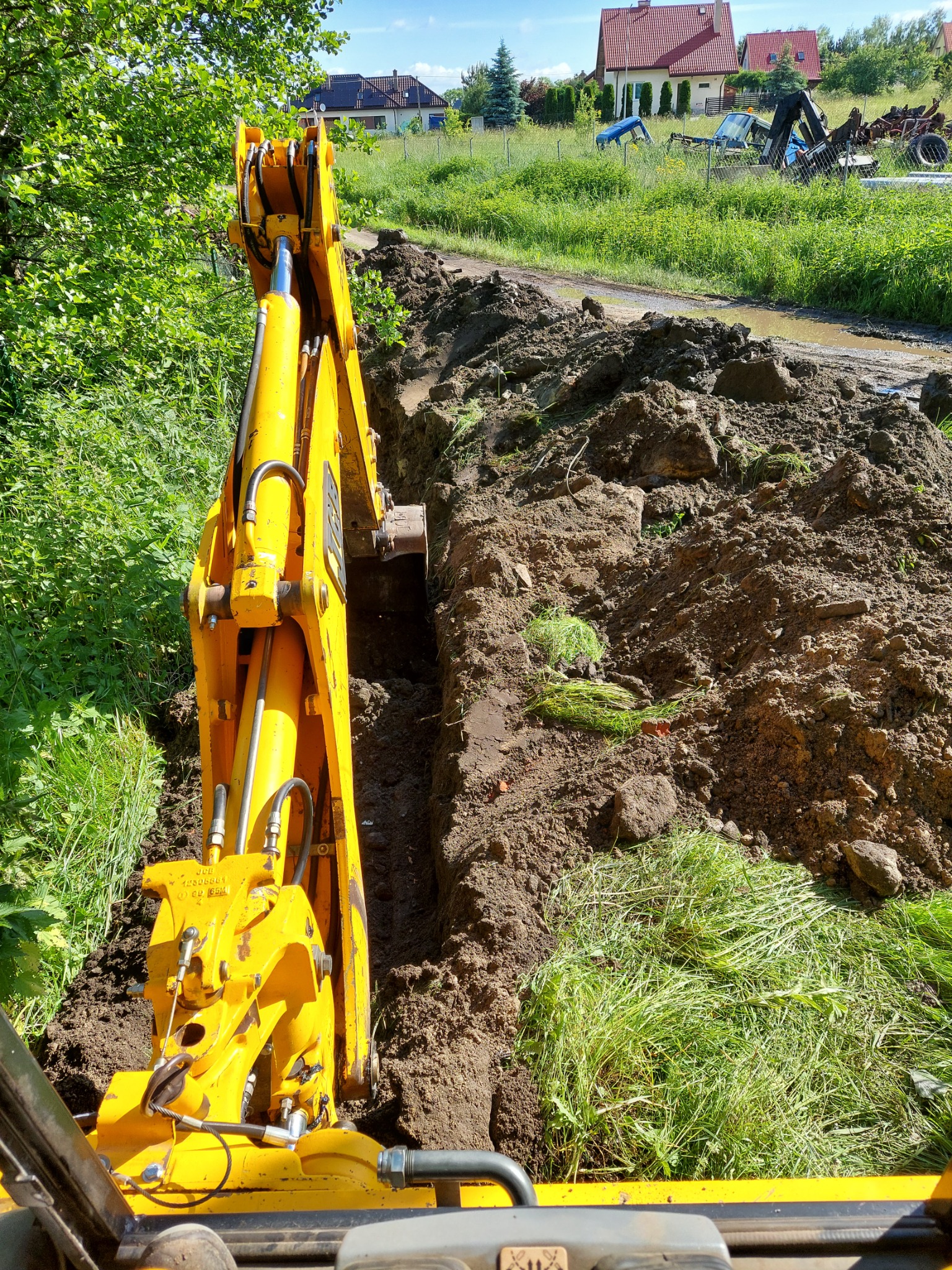 Żółta koparka podczas wykonywania wąskiego wykopu w ziemi, widok z perspektywy operatora, z fragmentami trawy i ziemi na krawędziach, w tle zabudowania wiejskie i stare maszyny rolnicze.