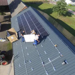 Montaż paneli słonecznych na metalowym dachu budynku gospodarczego; widoczne dwa busy, podnośnik teleskopowy dostarcza panele, częściowo zamontowane panele i elementy konstrukcji montażowej.