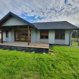 Nowoczesny dom z szarą elewacją, ciemną dachówką i drewnianymi elementami wykończeniowymi, widoczny z perspektywy trawnika w słoneczny dzień.