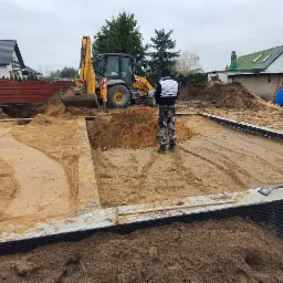 Przygotowanie terenu pod budowę domu: ubijanie piasku zagęszczarką, częściowo wykonane fundamenty z izolacją, koparka i hałdy ziemi w tle.
