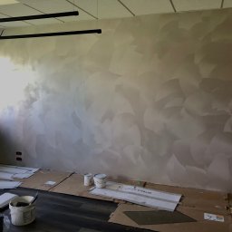 JACBIS Fotografia - Ściana pokryta dekoracyjną farbą w odcieniach beżu i szarości, z widoczną fakturą przypominającą pióra, zabezpieczona kartonem na podłodze, widoczne puszki z farbą i narzędzia malarskie.