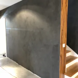 Ściana pokryta dekoracyjnym tynkiem w odcieniach szarości, z widoczną strukturą i efektem cieniowania, sąsiadująca z drewnianą kolumną i schodami.