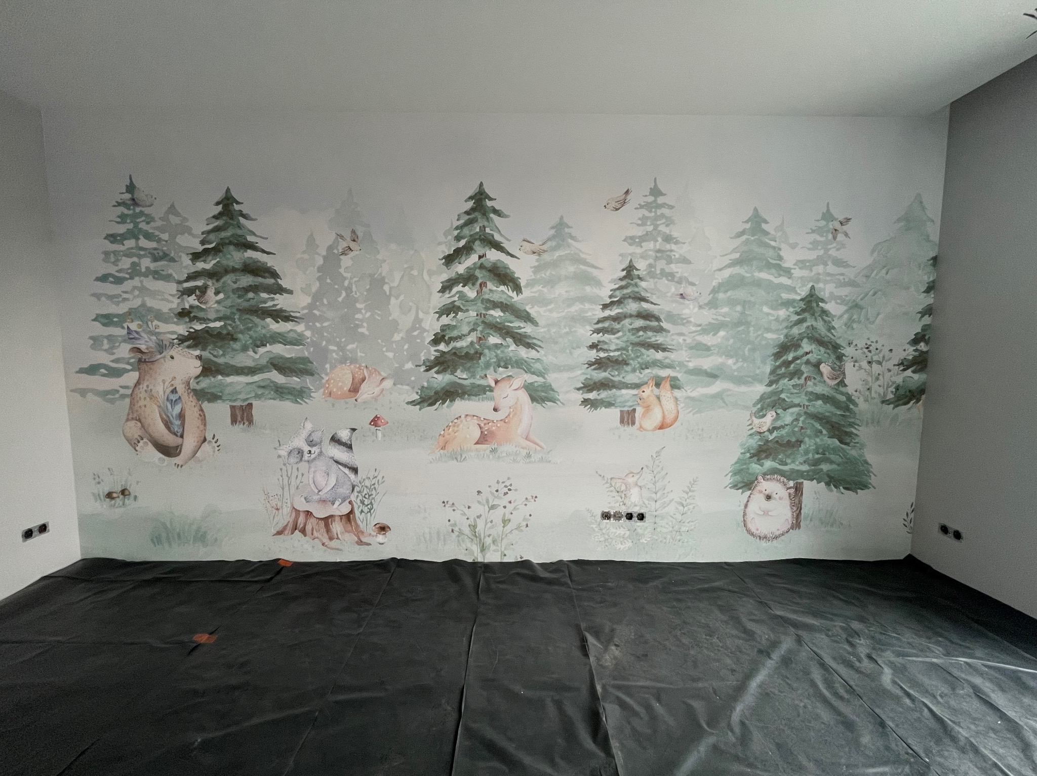 Ściana pokoju dziecięcego z tapetą przedstawiającą leśne zwierzęta i drzewa, zabezpieczona folią malarską na podłodze.