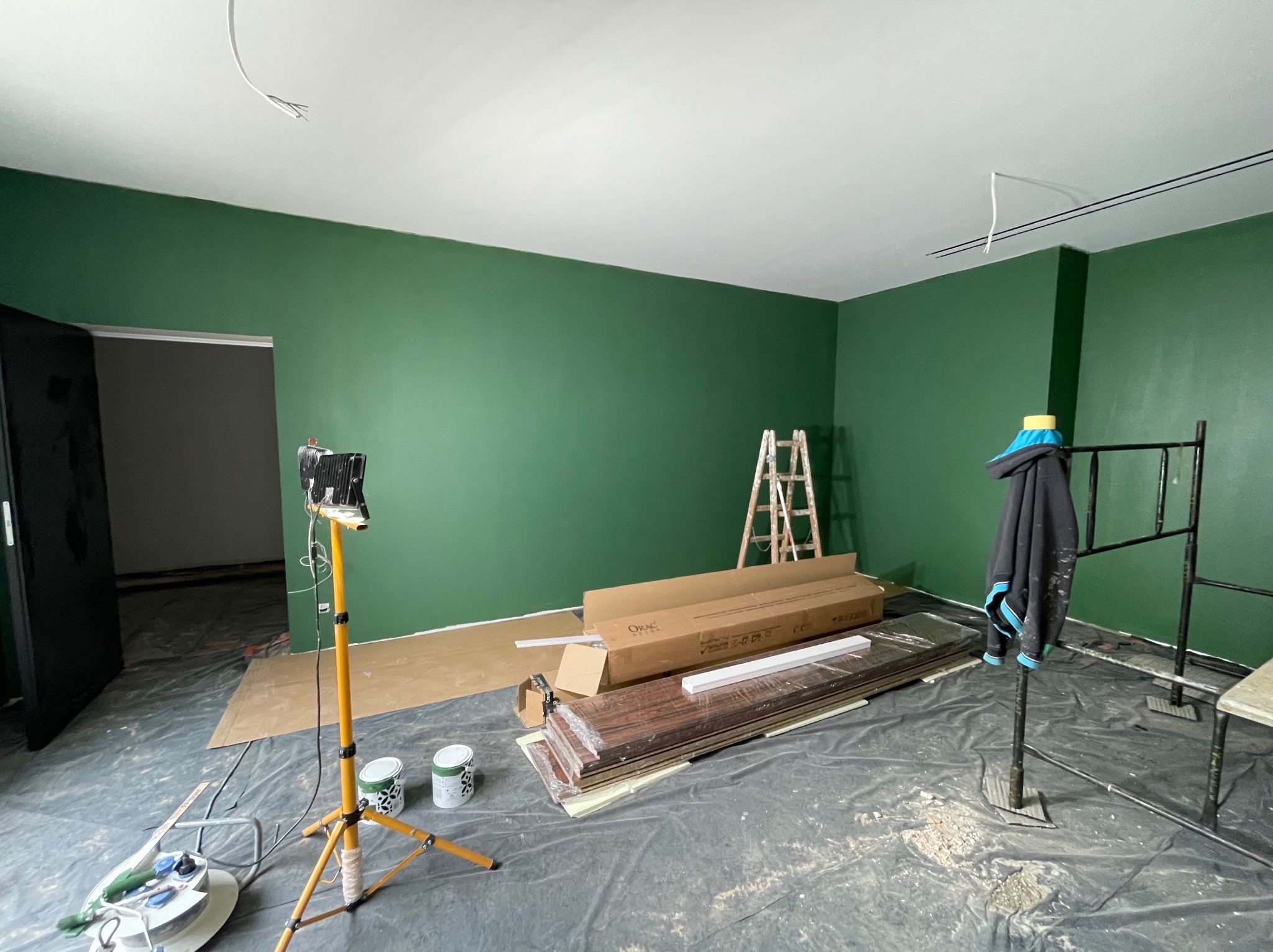 Pokój po malowaniu ścian na zielono, widoczne puszki z farbą, rozłożone panele podłogowe w opakowaniach, żółty statyw z lampą i drabina.