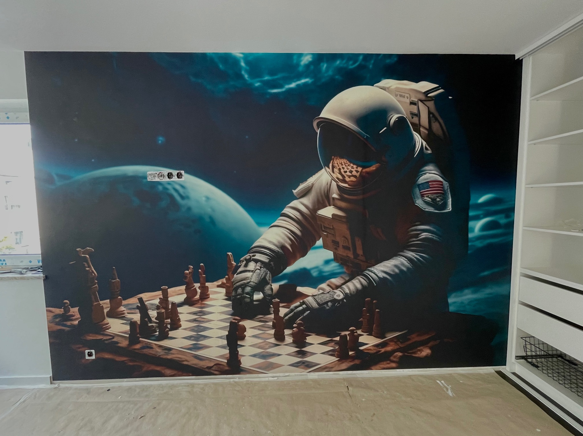 Tapeta na ścianie przedstawiająca astronautę grającego w szachy na tle kosmosu, widoczne gniazdka elektryczne i fragment szafy.