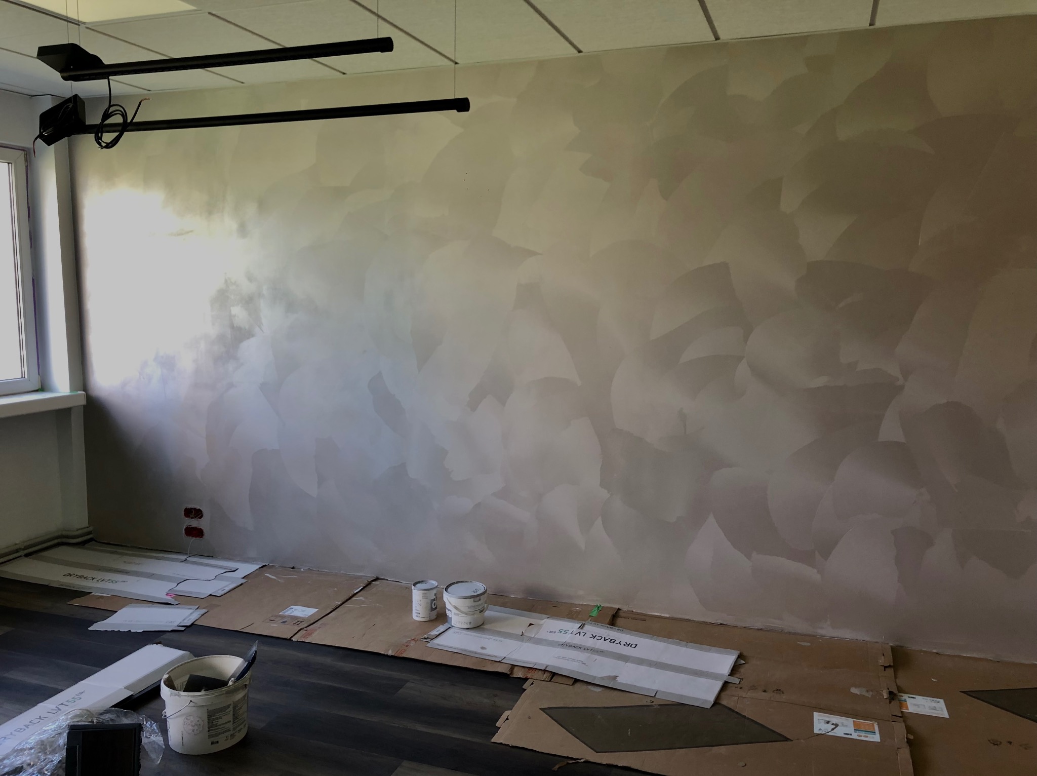 Ściana pokryta dekoracyjną farbą w odcieniach beżu i szarości, z widoczną fakturą przypominającą pióra, zabezpieczona kartonem na podłodze, widoczne puszki z farbą i narzędzia malarskie.