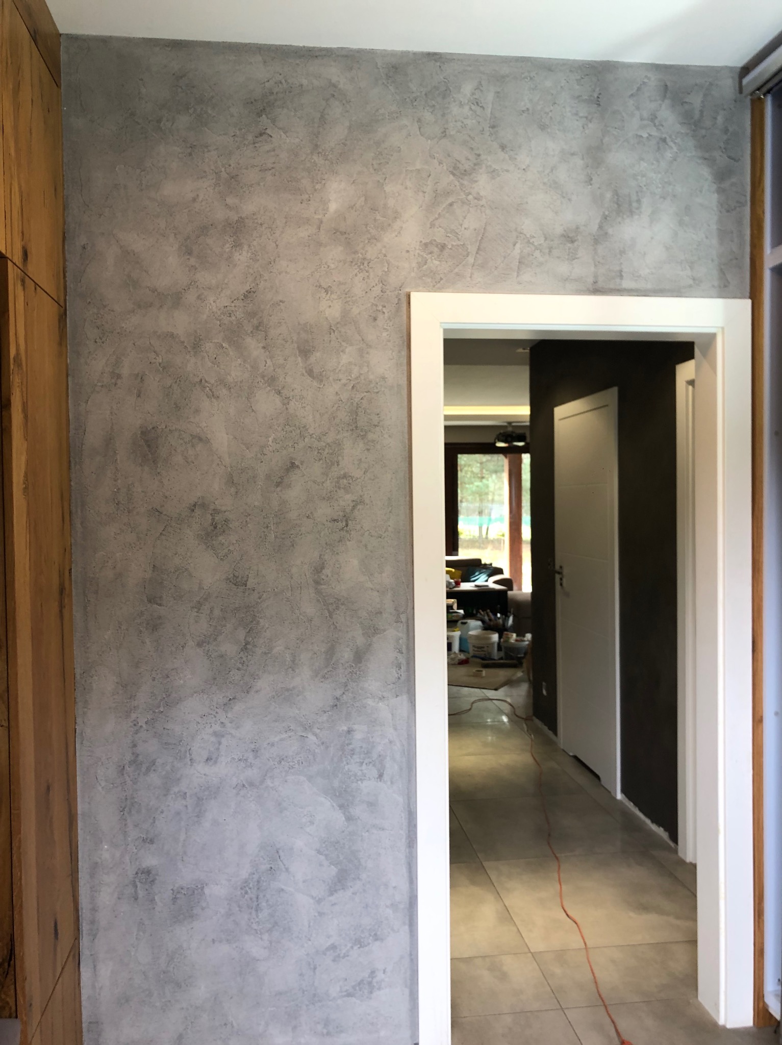 Szara ściana z efektem dekoracyjnym, widok na korytarz z białą ramą drzwiową, fragment drewnianej zabudowy po lewej stronie.