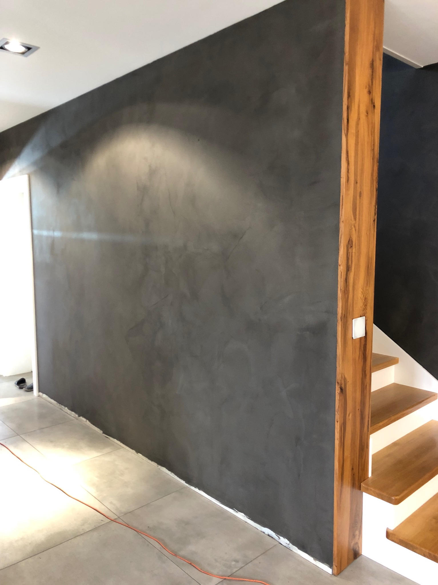 Ściana pokryta dekoracyjnym tynkiem w odcieniach szarości, z widoczną strukturą i efektem cieniowania, sąsiadująca z drewnianą kolumną i schodami.