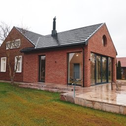 marcin pingwara architekt - Nowoczesny dom z cegły klinkierowej i dużymi przeszkleniami, widok z zewnątrz na wilgotny taras. W tle drugi budynek z panelami słonecznymi.