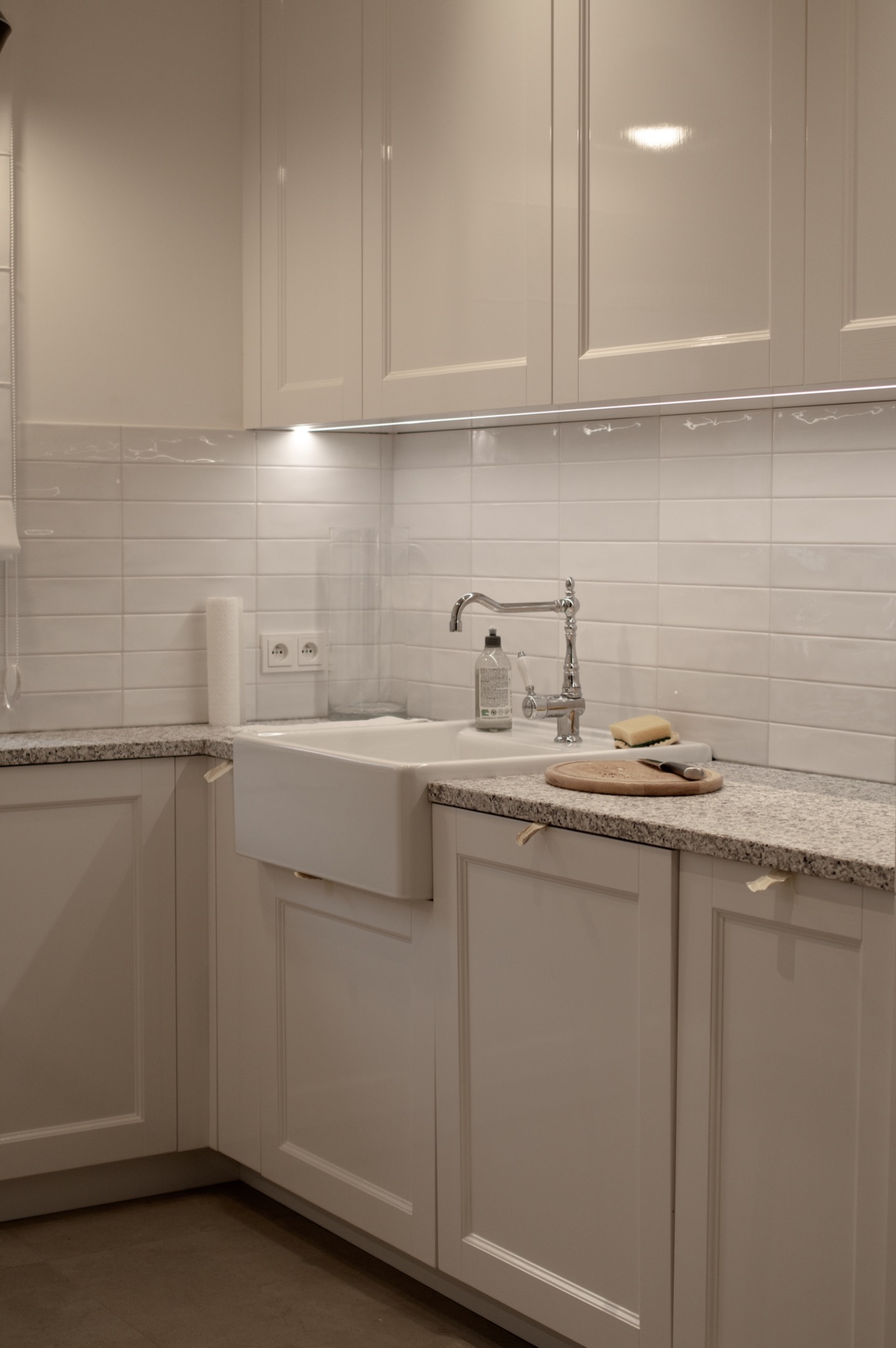 Jasna, klasyczna kuchnia z białymi szafkami, granitowym blatem, ceramicznym zlewem farmerskim i chromowaną baterią; na blacie deska do krojenia, nóż i gąbka.