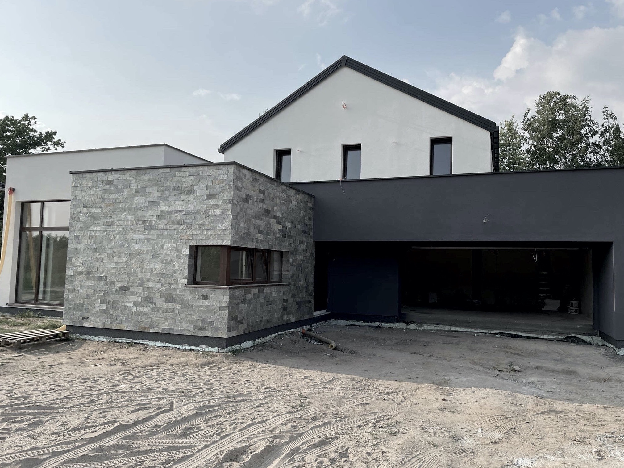 Nowoczesny dom jednorodzinny z elewacją z kamienia naturalnego i otwartym garażem, w trakcie wykańczania terenu wokół budynku. Widoczne ślady opon na piasku.