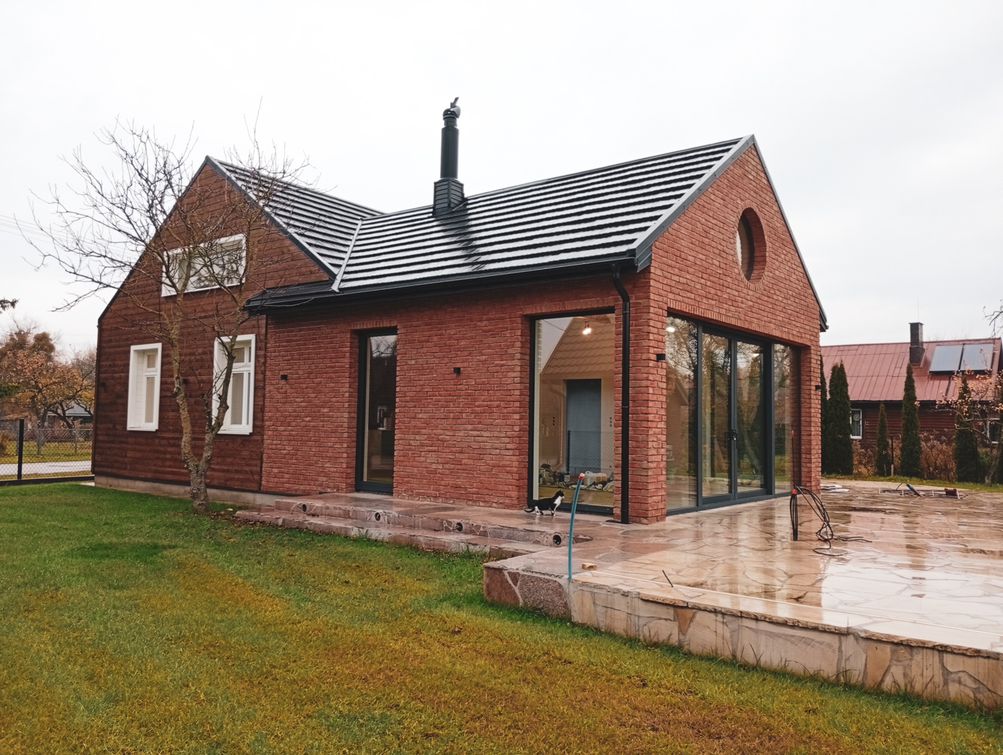 Nowoczesny dom z cegły klinkierowej i dużymi przeszkleniami, widok z zewnątrz na wilgotny taras. W tle drugi budynek z panelami słonecznymi.